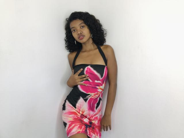 CandyMagicX, with curly black hair, wearing a strapless black dress with pink floral patterns, touches her face, showing off her small ass and dark brown skin.