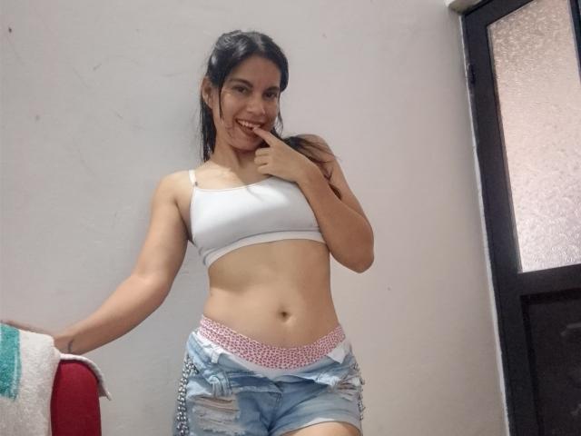 EstrellaFandy with dark hair, smiling, finger in mouth, wearing white sports bra, showing small tits, and blue ripped shorts with pink waistband, standing in a simple room.
