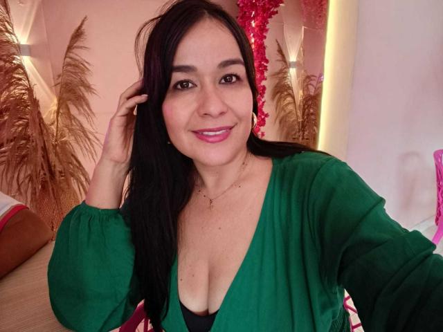 TamaraFoxy con capelli neri, vestito verde, rossetto rosa, che sorride, toccandosi i capelli, mostrando il seno, collana d'oro, decorazioni rosa e dorate, luce rosa tenue sullo sfondo..