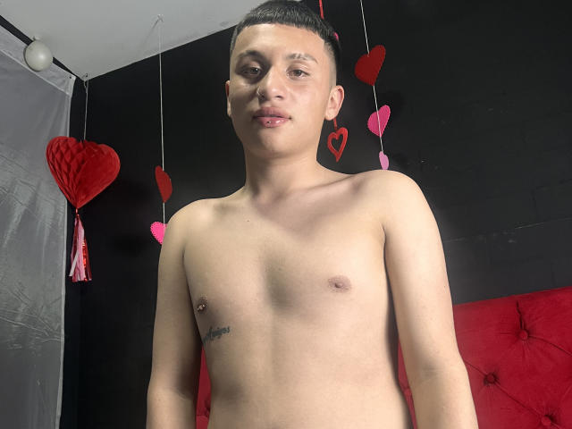 AronSweety, shirtless, standing with a slight smile, small nipple piercings, short dark hair, light skin, tattoo on right side, red and pink heart decorations in background.