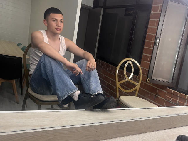 AronSweety, short-haired dude with light skin, white tank top, blue jeans, black socks, and black shoes, sitting on a chair, arms resting on knees, looking at camera.