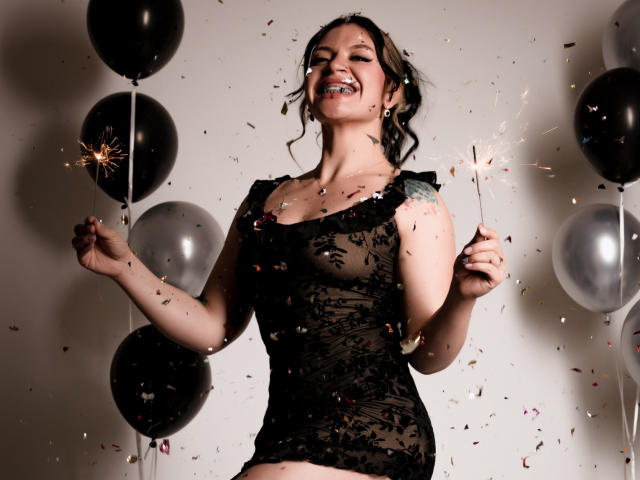 KatalinaJohnson, souriante, tenant des étincelles, robe en dentelle noire, confettis, ballons noirs et argentés, cheveux bouclés foncés, tatouage visible sur l'épaule droite, célébrant, plan moyen, debout..