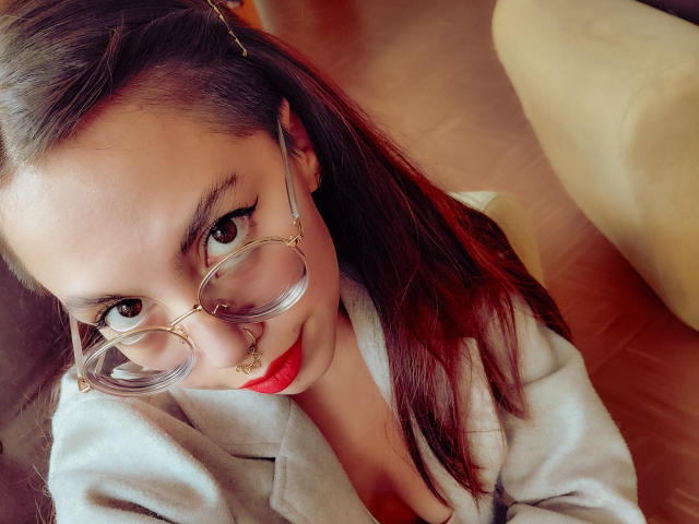 NaturalMariaHun met rode lippenstift, heldere brillen en een neusring, die omhoog kijkt met donkerbruine ogen, een witte blazer draagt en bruin haar heeft met rode highlights..