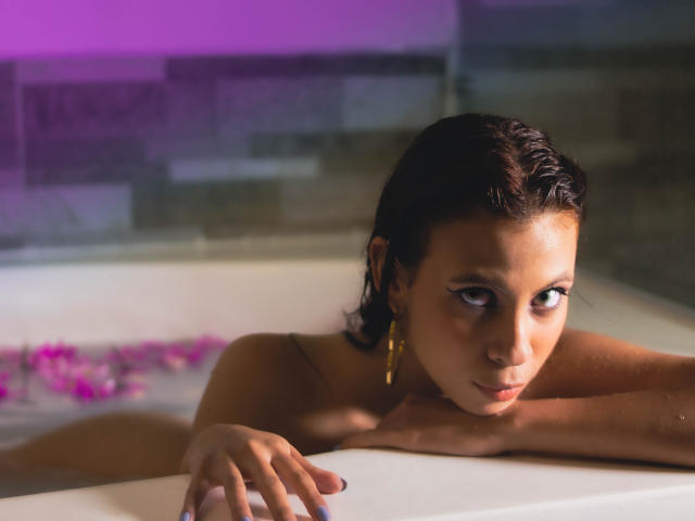 ElevenSkys with wet brown hair, looking down, arms resting on white bathtub edge, small gold earrings, purple and gray background, purple light, purple flowers in water.