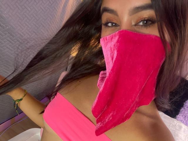 EmyXCute, con cabello largo y oscuro, vistiendo una máscara de terciopelo rosa que cubre su nariz y boca, un top de bikini rosa con tirantes, una pulsera verde, y mirando a la cámara con ojos marrones..