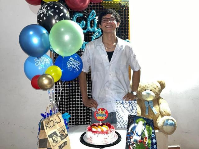 SamuelHills, met kort zwart haar, een bril en een wit shirt, staat lachend achter een tafel met een taart, een teddybeer en ballonnen, en houdt een kaart vast..