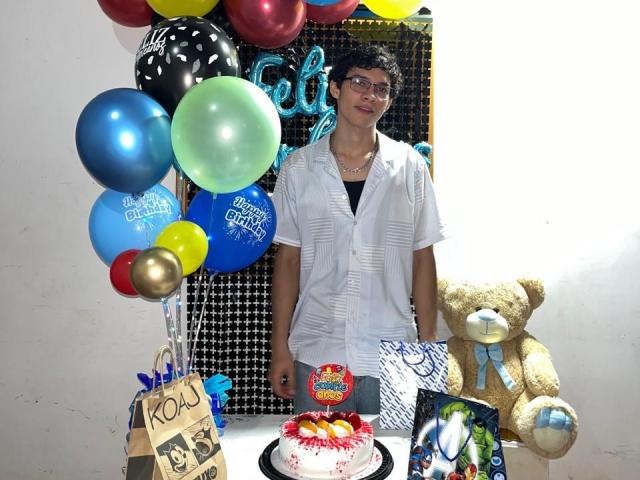 SamuelHills, short-haired dude with glasses, wearing a white shirt, standing next to a table with a cake, teddy bear, balloons, and a card. Cake has red and white frosting.