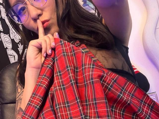 EviieGothel with red plaid shirt, black bra, sheer top, finger on lips, glasses, and tattoo on left arm, looking seductive. Boobs partially visible. EviieGothel's hair is brown, long.