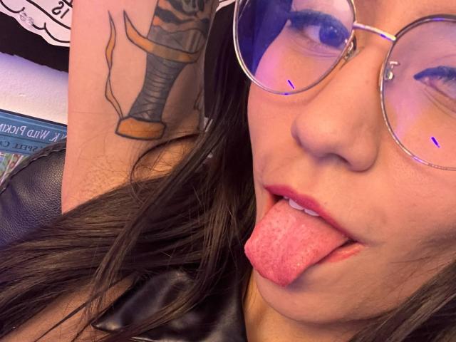 EviieGothel with round glasses, sticking out pink tongue, showing a tattoo of a boot on her arm, wearing a black leather top. Her face is close to the camera.