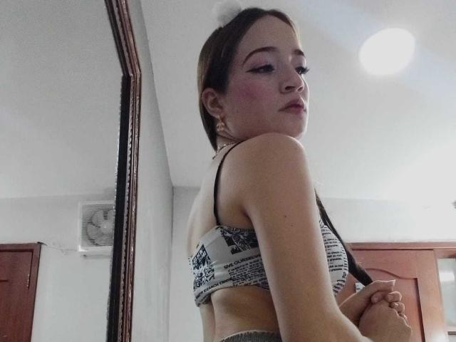 MadisonAngel con un leggero rossore sulle guance, capelli castani raccolti in una coda di cavallo, indossa un reggiseno bianco e nero, guarda oltre la spalla, tiene un braccio, è in piedi di fronte a uno specchio..