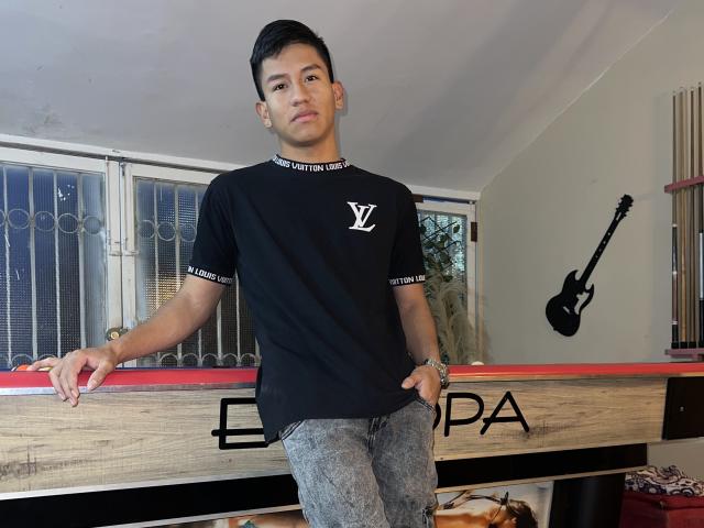 SexInTheBar with short black hair, wearing a black tee with a white "V" logo and grey jeans, leaning on a wooden table with a guitar on the wall.