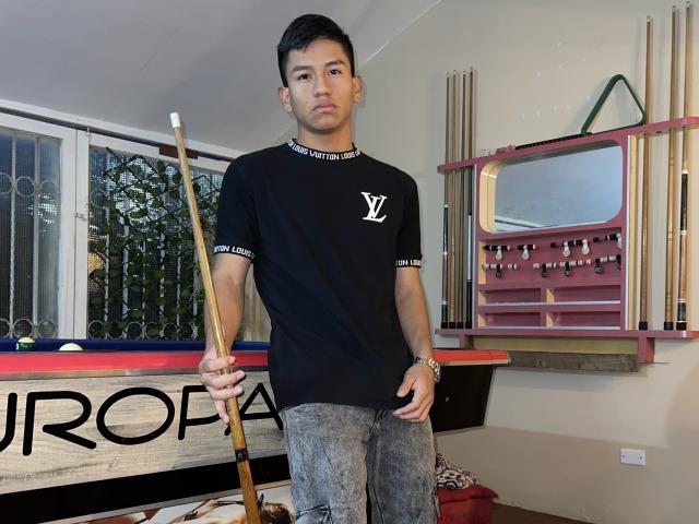 SexInTheBar stands holding a pool cue, wearing a black t-shirt with a white logo, and gray jeans, with a serious expression, in a room with a pool table.