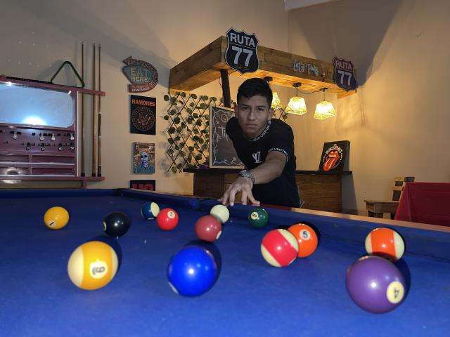 SexInTheBar, a young dude with short black hair, wearing a black shirt, points at colorful pool balls on a blue felt table, looking focused, in a dimly lit room.