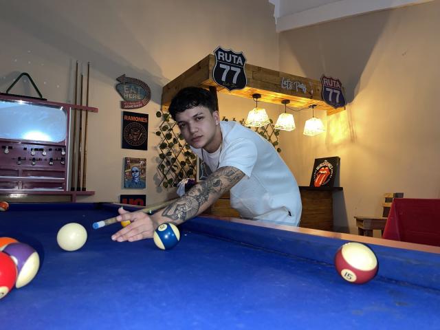 SexInTheBar with tattoos on right arm, wearing white shirt, leaning over blue pool table, aiming at ball with pool cue, surrounded by colorful pool balls.