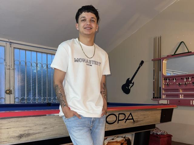 SexInTheBar with short curly dark hair, tattoos on both arms, white "MOMAARTIST" t-shirt, gold necklace, blue jeans, hands in pockets, standing by pool table.