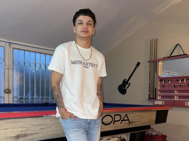 SexInTheBar with short dark hair, wearing a white t-shirt, blue jeans, and a necklace, standing in front of a pool table, showing tattoos on both arms, hands in pockets.