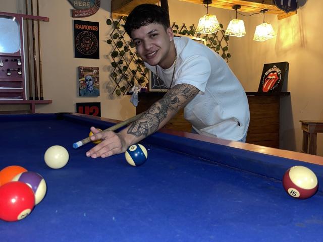 SexInTheBar with tattoos on his arm, wearing a white shirt, leaning over a pool table, aiming a cue stick at a ball, smiling, with various pool balls on the table.