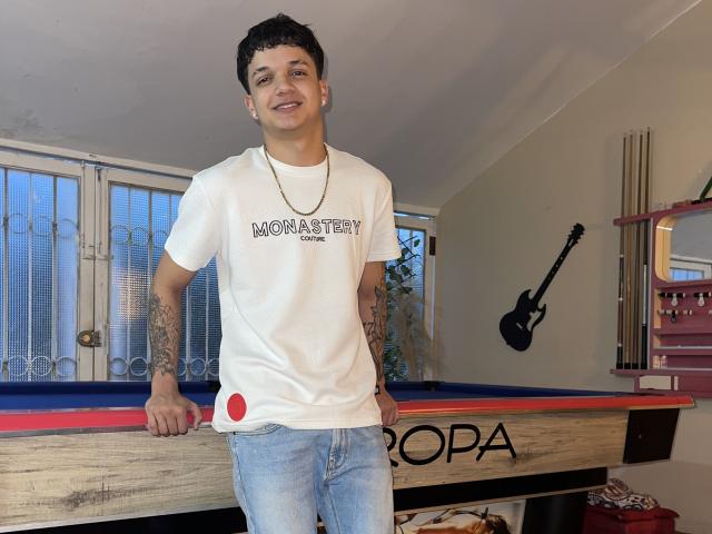 SexInTheBar with short dark hair, tattoos on left arm, white t-shirt, blue jeans, necklace, leaning on pool table, smiling, holding red pool ball, standing in well-lit room.