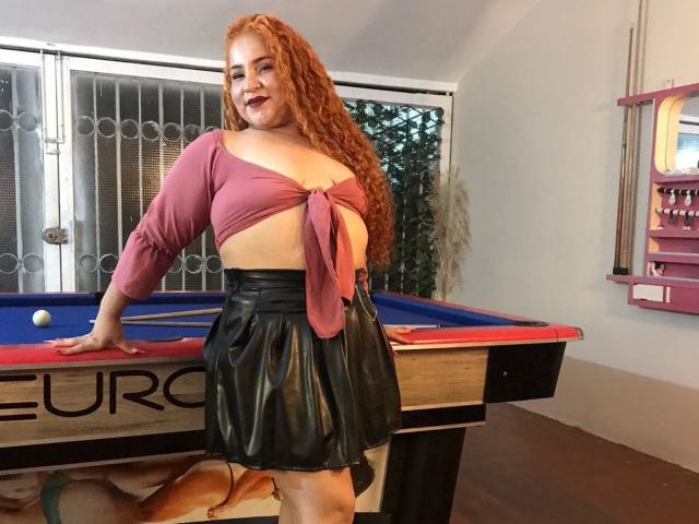 Redhead SexInTheBar with curly hair, pink tied top showing her boobs, black leather skirt, leaning on pool table, smiling, white walls, glass doors, and pink shelf in background.