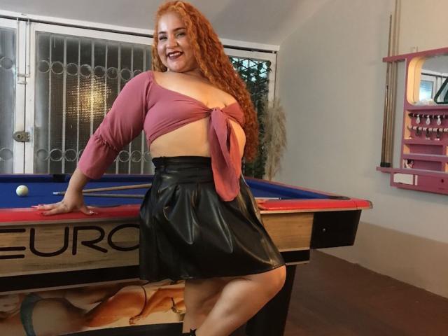 Sexy red-haired woman with curly hair, pink top tied below boobs, black leather skirt, smiling, leaning on pool table, one hand on table, pool ball visible, pink wall.