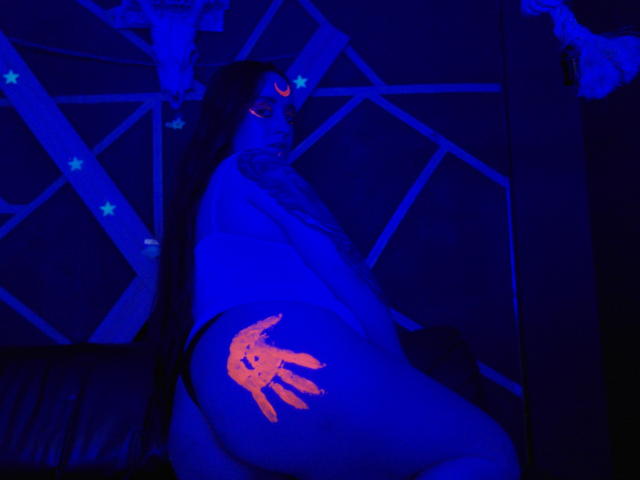 GelenaBlack with glowing orange hand on ass, blue neon lighting, long dark hair, tattoo on upper arm, skull decorations, leather couch, crescent moon on face, intense blue glow.