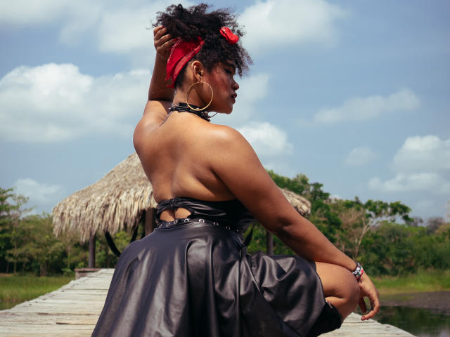 AliceJelanni with curly black hair, red headband, hoop earrings, black leather dress, showing her back, sitting on wooden dock, hand in hair, blue sky, white clouds.