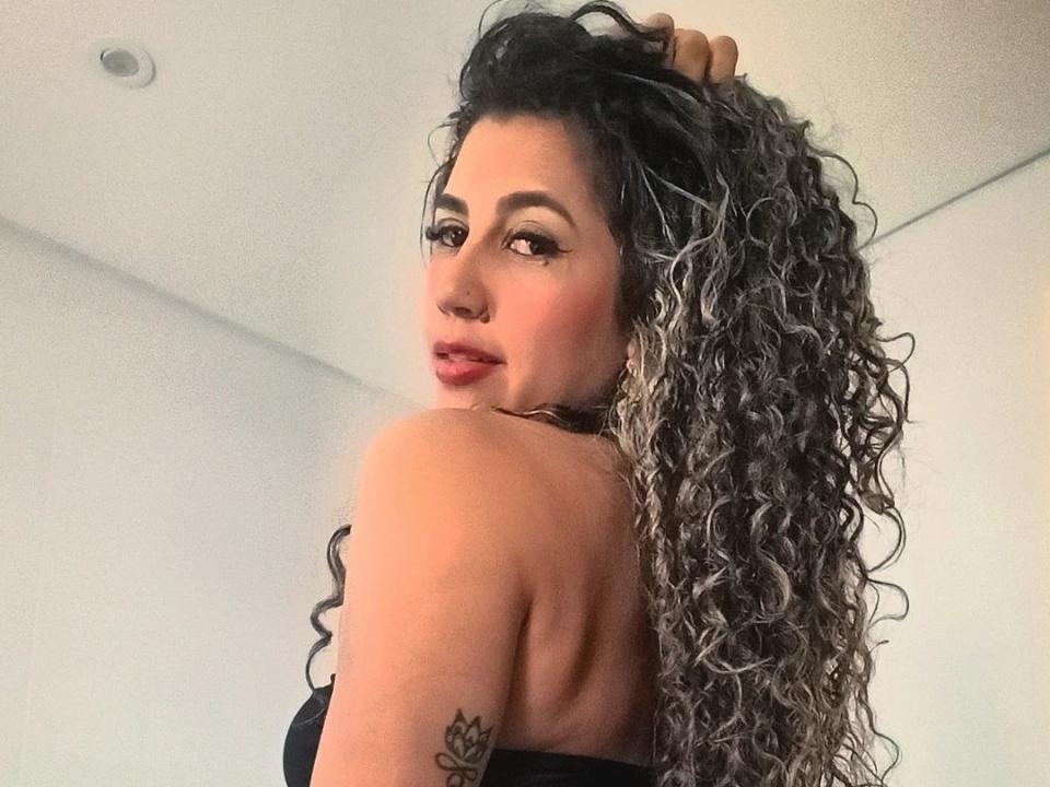 LilyCorner1 with long, curly, gray-to-black hair, holding it up, looking over her shoulder, showing her tattooed arm, and black strapless top, with a slight smile.