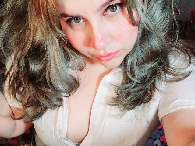 Akkanee with green eyes, wavy grayish-brown hair, and pinkish skin, wearing a white button-up shirt, pouting her pink lips, showing her small tits.