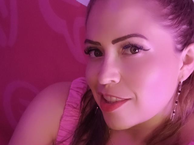 FantastykLadies has pink lipstick, winged eyeliner, and brown eyes, smiling with red-pink hair, wearing a pink ruffled top, and pink earrings, under purple lighting.