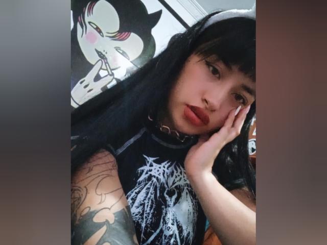 Lxlitx with black hair, red lipstick, and tattoos, touching her face, wearing a black graphic tee, looking at the camera, with a cat-ear headband.