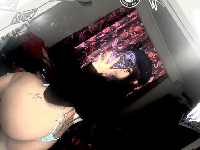 Lxlitx with a black beanie, phone covering face, wearing a black top and light blue thong, showing a tattoo on her left butt cheek and a tattoo on her right side.
