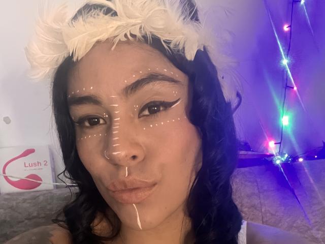 StellaBeckker has white lines on her face, black wavy hair, a white feather headband, and a nose ring, with a small drop of white fluid on her chin.