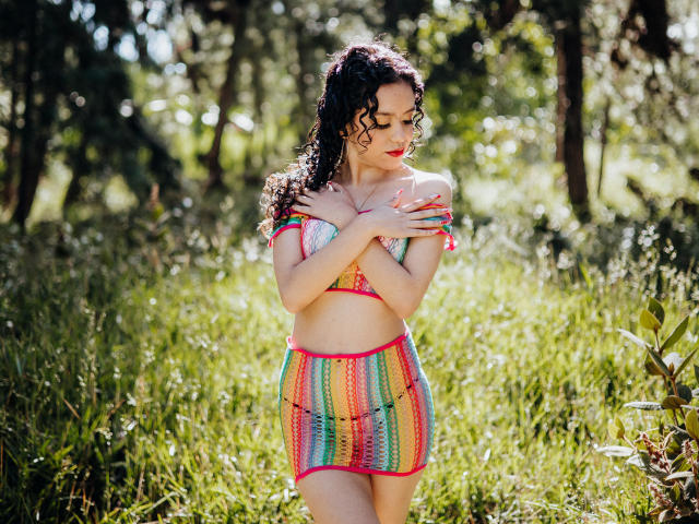 Macaroux with curly black hair, red lipstick, and colorful crochet outfit, covering her small tits with hands, standing in sunlit grassy area, looking down.
