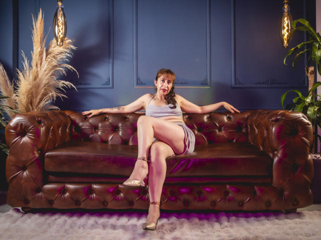 ViktoriaBrown with tits and ass in a gray dress, sitting on a brown leather couch, legs crossed, arms out, gold heels, puffy brown plant, dark blue wall, and wall sconces.