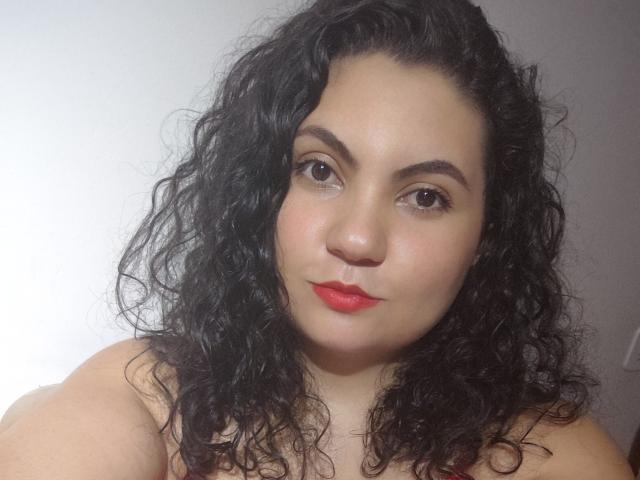 Marymanroe has curly black hair, red lipstick, and brown eyes, with a fair skin tone, and is looking directly at the camera with a slight smile.