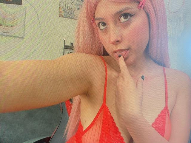 CatalinaRogers met roze haar, een rode kantbeha, een vinger in haar mond, die haar borsten laat zien, heeft een roze haarspeldje, zwarte nagellak en een sexy uitstraling op een selfie..