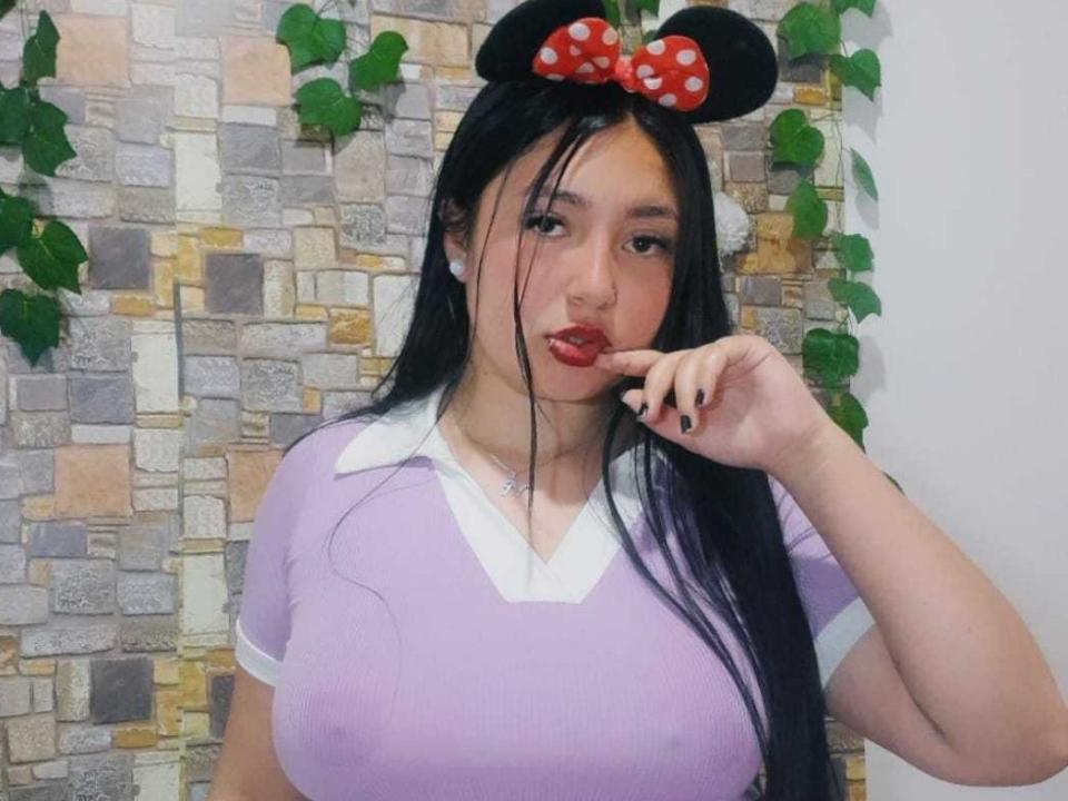NikkyCuper, con i capelli neri, le labbra rosse, indossa una camicia viola, un colletto bianco e orecchie da Minnie Mouse con un fiocco rosso a pois, mentre tocca le labbra. Il suo seno è visibile..