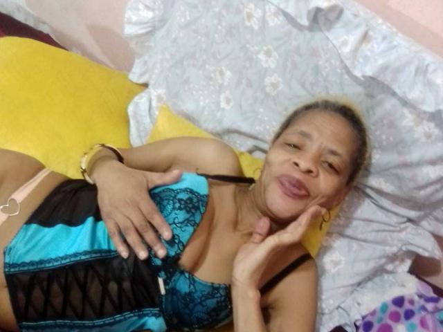 LongSexyNipples, deitada na cama, vestindo lingerie de renda azul e preta, lábios cheios, mão no peito, travesseiro amarelo, pulseira dourada, cabelo loiro, seios pequenos, sexy, confiante..