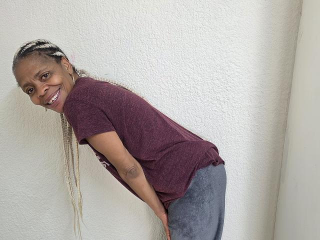 LongSexyNipples with dark brown skin, smiling, wearing a maroon tee and grey pants, bending forward, long blonde braids, hands on butt, white textured wall in background.
