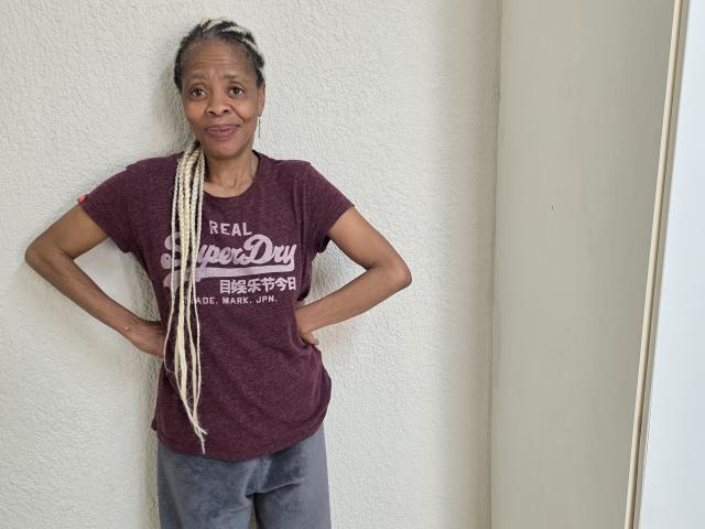 LongSexyNipples se tient les mains sur les hanches, portant un t-shirt marron "Real Hero Day" et un pantalon gris, avec de longues tresses blanches et un petit sourire, dans une pose décontractée..