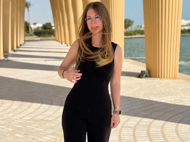 Soleea-hot with long brown hair, wearing a tight black sleeveless top and black pants, standing under tall yellow columns, sporting pink glasses and a black wristwatch.