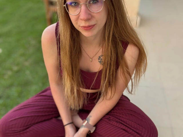 Soleea-hot with long brown hair, glasses, and a small tattoo, sitting cross-legged in maroon clothes, showing her boobs slightly, and a cute smile, hands on knees.