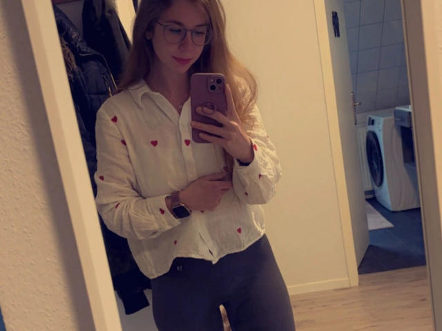 Soleea, bellissima con lunghi capelli castani, occhiali, camicia bianca con cuori rossi, pantaloni grigi, che tiene in mano un telefono, scatta un selfie allo specchio, indossa un orologio, sorride leggermente, in una stanza con della biancheria..