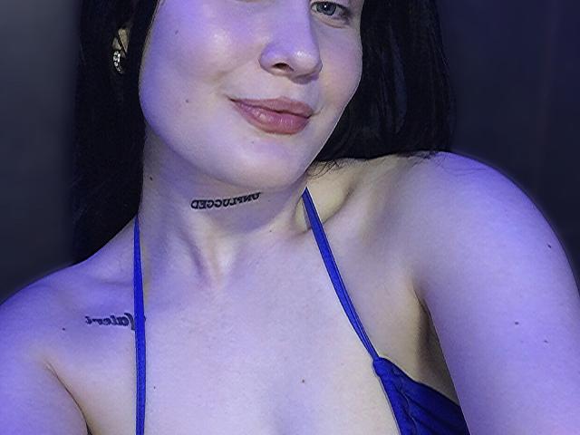 BluesPatty with black hair, pink shirt, and dark blue bra, standing by a lake, hand in hair, showing small tits, cute and calm, pink shirt buttoned halfway.