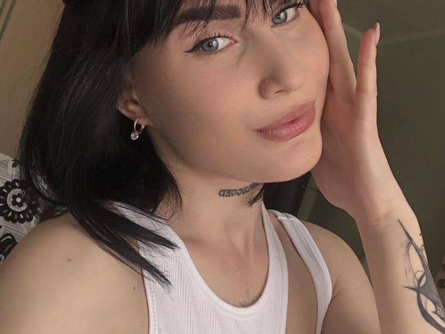 BluesPatty, con capelli neri, occhiali, reggiseno sportivo nero e tatuaggio sul braccio destro, che parla al telefono, seduta vicino a un lago verde, tenendo il telefono all'orecchio destro, guardando di lato..