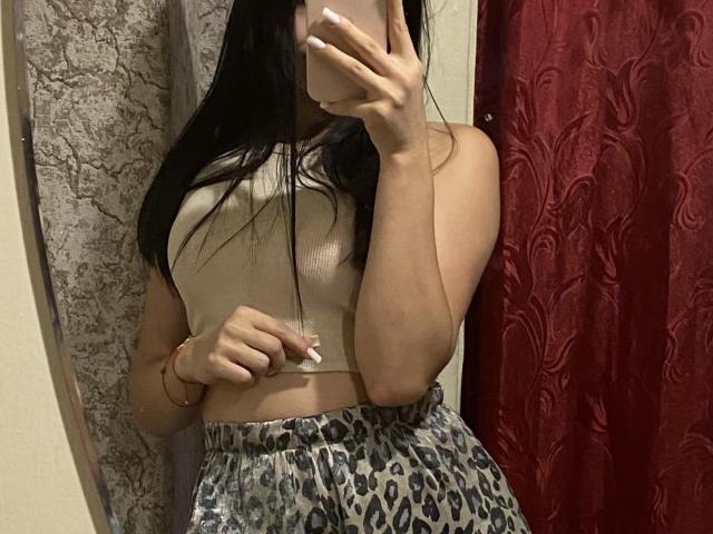 Jaurita, con la pelle marrone scuro, i capelli lunghi e intrecciati, indossa lingerie di pizzo nera, è seduta con le gambe divaricate, sorride, ha una mano tra i capelli, piccoli seni, è carina, sexy, e indossa una collana e un braccialetto..