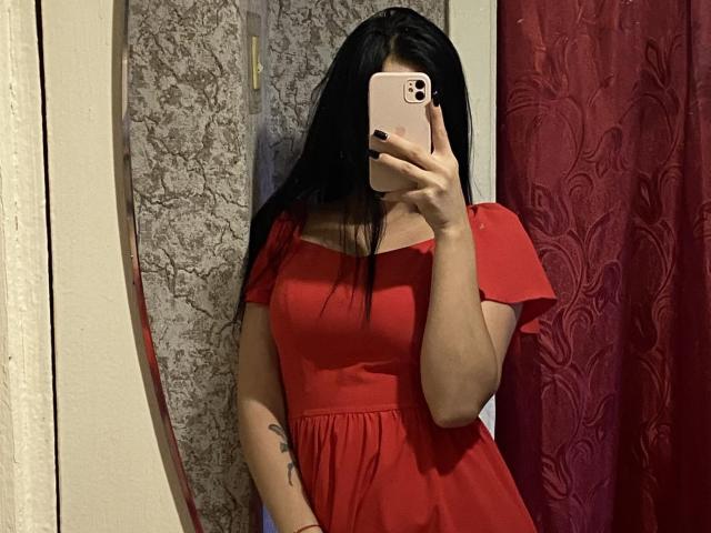 FortuneElizabeth with long black hair, holding phone, wearing a sheer beige top showing small tits, and leopard print skirt, standing in front of a red curtain.