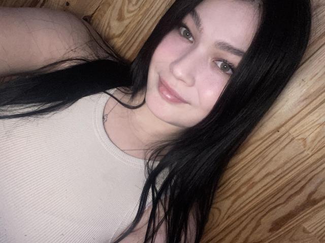 FortuneElizabeth tem cabelos pretos longos, pele clara, olhos verdes e um sorriso discreto com lábios rosados. Ela está vestindo uma camisa branca com uma alça preta..