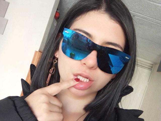 FortuneElizabeth with long black hair, holding pink roses in front of her bare tits, taking a mirror selfie with a pink phone, wearing a black strap top and silver bracelet.