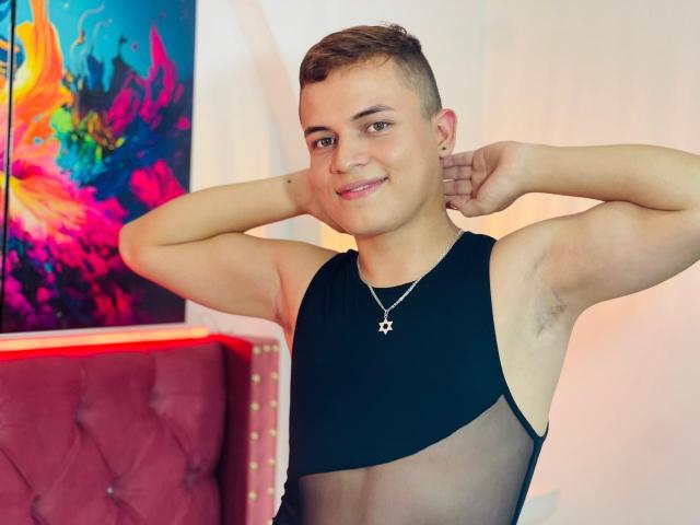 LeonAdams, young dude with short brown hair, smiling, arms flexed, wearing black sleeveless top with sheer chest, star necklace, armpit hair visible, standing next to red velvet chair.